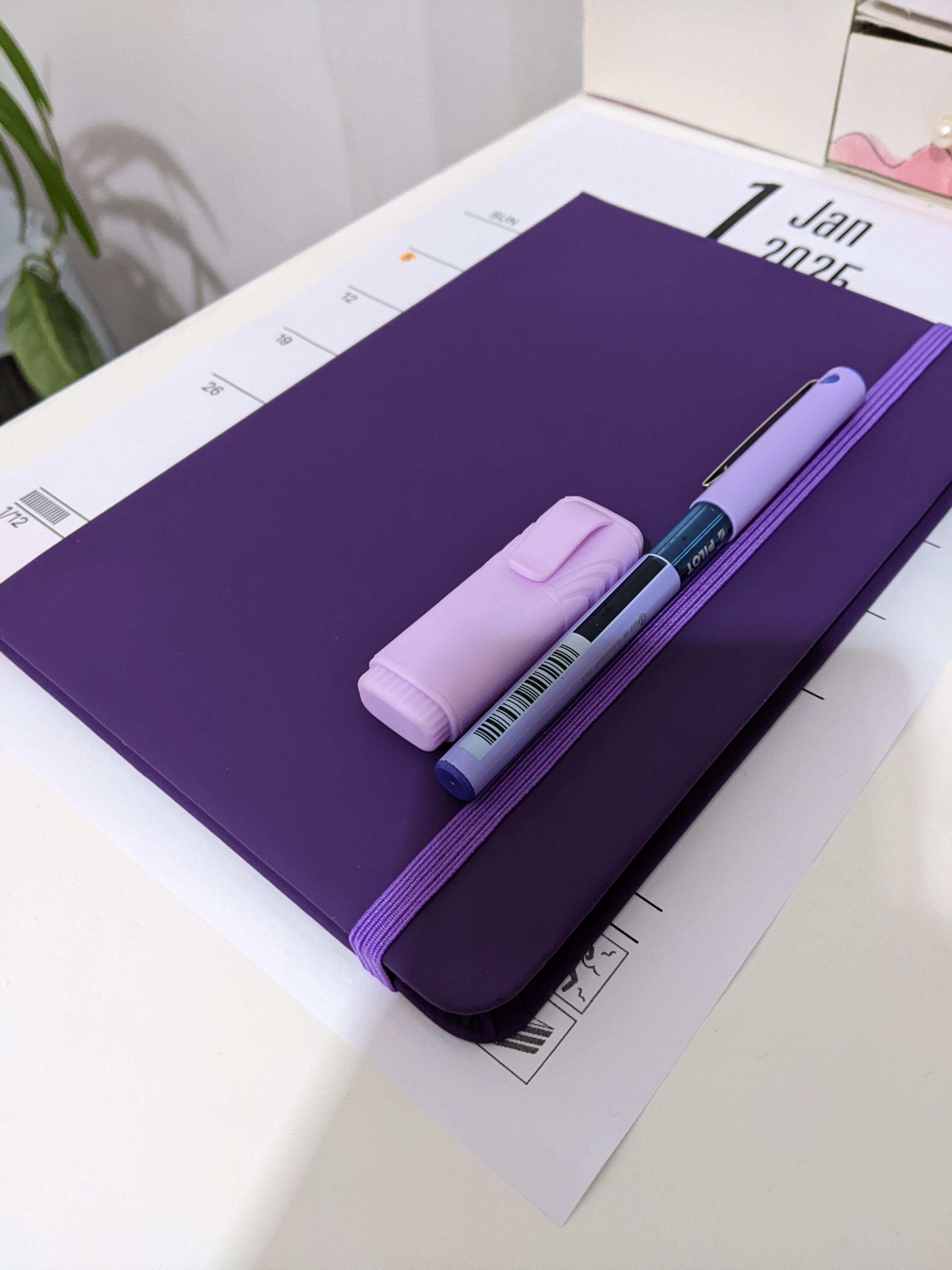 Smooth, “folded in half A4 sheet” sized journal, on top of which there’s a purple gel pen and a short (half the length of a normal one) purple highlighter.